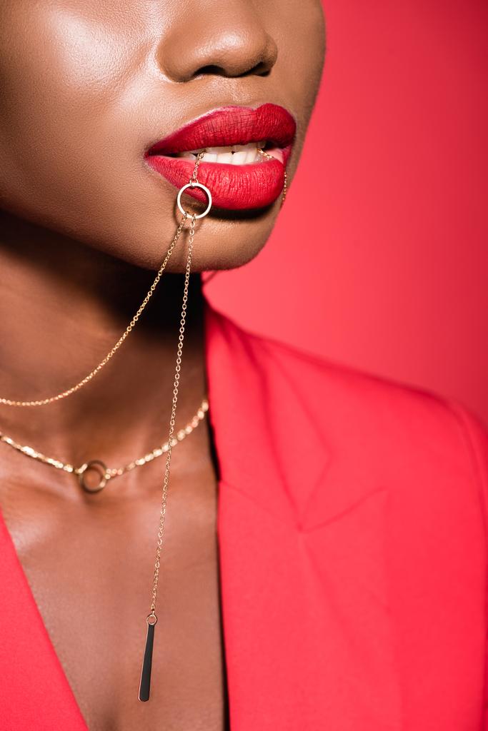 vista cortada de Africano americano jovem mulher na roupa elegante segurando colar na boca isolado no vermelho - Foto, Imagem