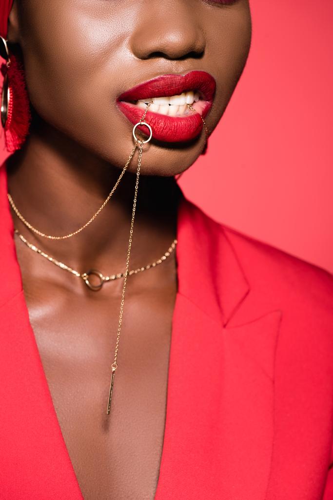 обрезанный вид на африканскую американскую молодую женщину в стильном наряде держа ожерелье во рту изолированы на красный - Фото, изображение