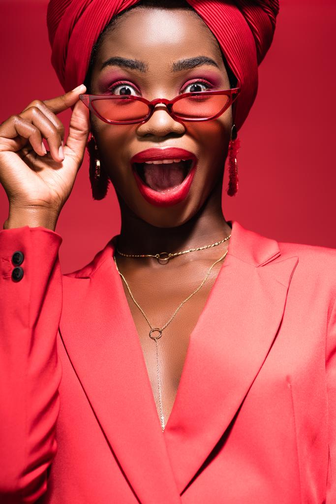 aufgeregten afrikanisch-amerikanischen junge Frau in stilvollem Outfit, Sonnenbrille und Turban isoliert auf rot - Foto, Bild
