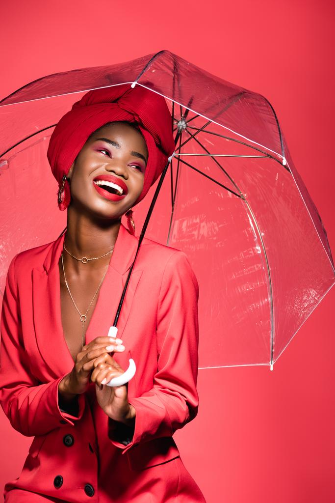 glücklich afrikanisch-amerikanische junge Frau in stilvollem Outfit und Turban hält Regenschirm isoliert auf rot - Foto, Bild