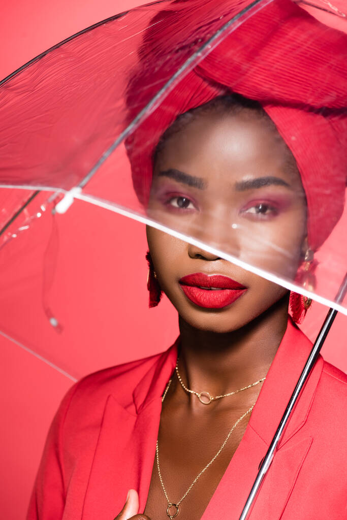 africká americká mladá žena ve stylovém oblečení a turban držení deštník izolované na červené - Fotografie, Obrázek
