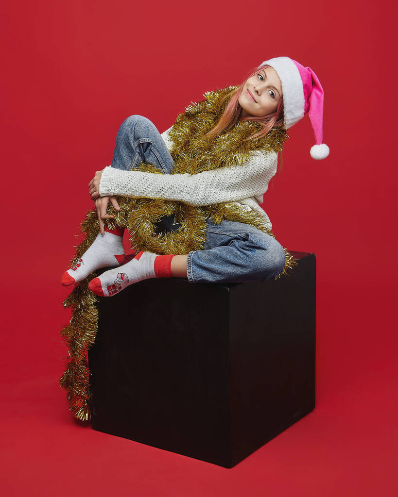 サンタの帽子の少女が黒い立方体に座っている。赤を背景にしたスタジオでの撮影会 - 写真・画像