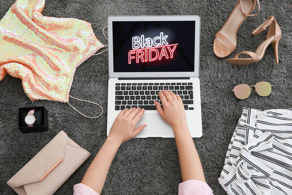 Woman surrounded by clothes and accessories using laptop with Black Friday announcement on grey carpet, top view - Photo, Image