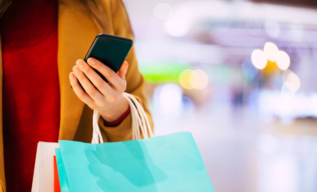 Feche a foto de mãos femininas com saco de compras e telefone inteligente no shopping enquanto compra algumas roupas - Foto, Imagem
