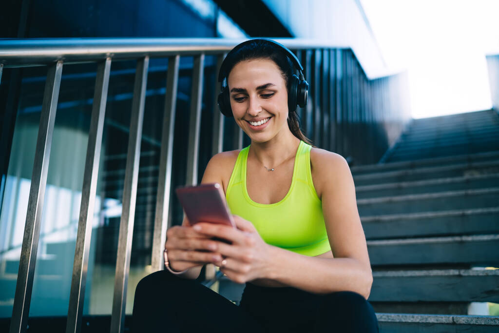 Lage hoek van optimistische vrouwelijke atleet in casual sportkleding zittend op betonnen trap en met behulp van smartphone en koptelefoon rusten na harde training - Foto, afbeelding