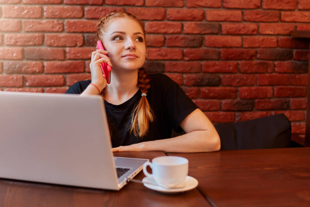 Αξιολάτρευτη γυναίκα που μιλάει στο smartphone στο cafe ενώ εργάζεται στο διαδίκτυο μέσω φορητού υπολογιστή, κυρία τζίντζερ με σκεπτική έκφραση προσώπου που κάθεται στον καναπέ ενάντια σε τοίχο από τούβλα, γυναίκα που φοράει μαύρο casual t πουκάμισο. - Φωτογραφία, εικόνα