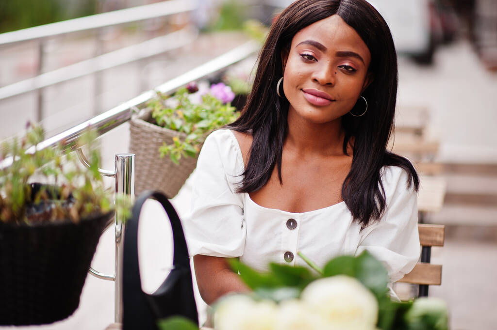 Όμορφη Αφροαμερικανή κοπέλα που κρατά μπουκέτο με λευκά τριαντάφυλλα λουλούδια κατά τη χρονολόγηση στην πόλη. Μαύρη επιχειρηματίας με πολλά λουλούδια δίπλα στο τραπέζι σε υπαίθριο καφέ. - Φωτογραφία, εικόνα