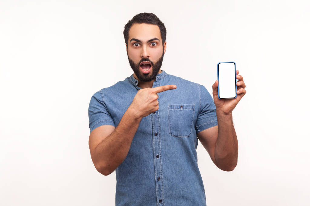 Šokovaný užaslý muž v modré košili ukazoval prstem na smartphone s prázdnou bílou obrazovkou a díval se na kameru s otevřenými ústy a velkýma očima. Vnitřní studio záběr izolované na bílém pozadí - Fotografie, Obrázek