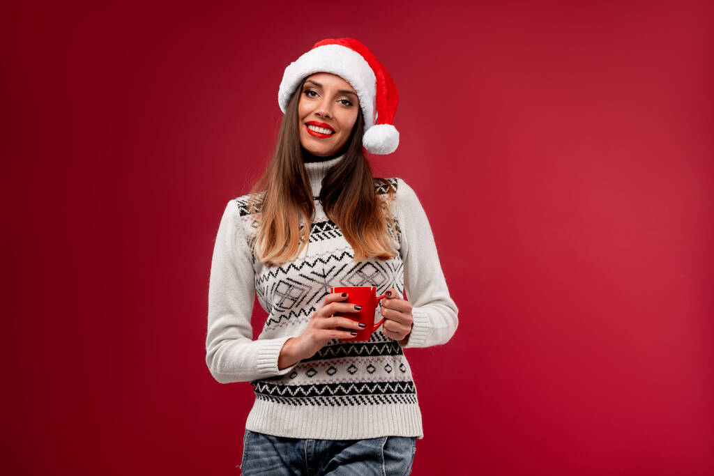 Zamknij portret beautifiul kaukaska kobieta w czerwonym kapeluszu Mikołaja na czerwonym tle studio. Boże Narodzenie Koncepcja Nowy Rok wakacje. słodkie dziewczyna zęby uśmiech pozytywne emocje z czerwony filiżanka kawy w dłoniach - Zdjęcie, obraz