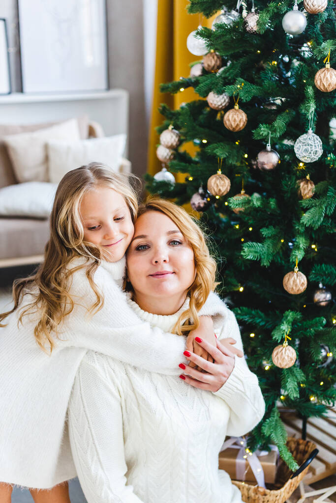 Ένα κορίτσι με τη μητέρα της κοντά στο χριστουγεννιάτικο δέντρο, το εσωτερικό διακοσμημένο για το νέο έτος και τα Χριστούγεννα, την οικογένεια και τη χαρά, τις παραδόσεις - Φωτογραφία, εικόνα