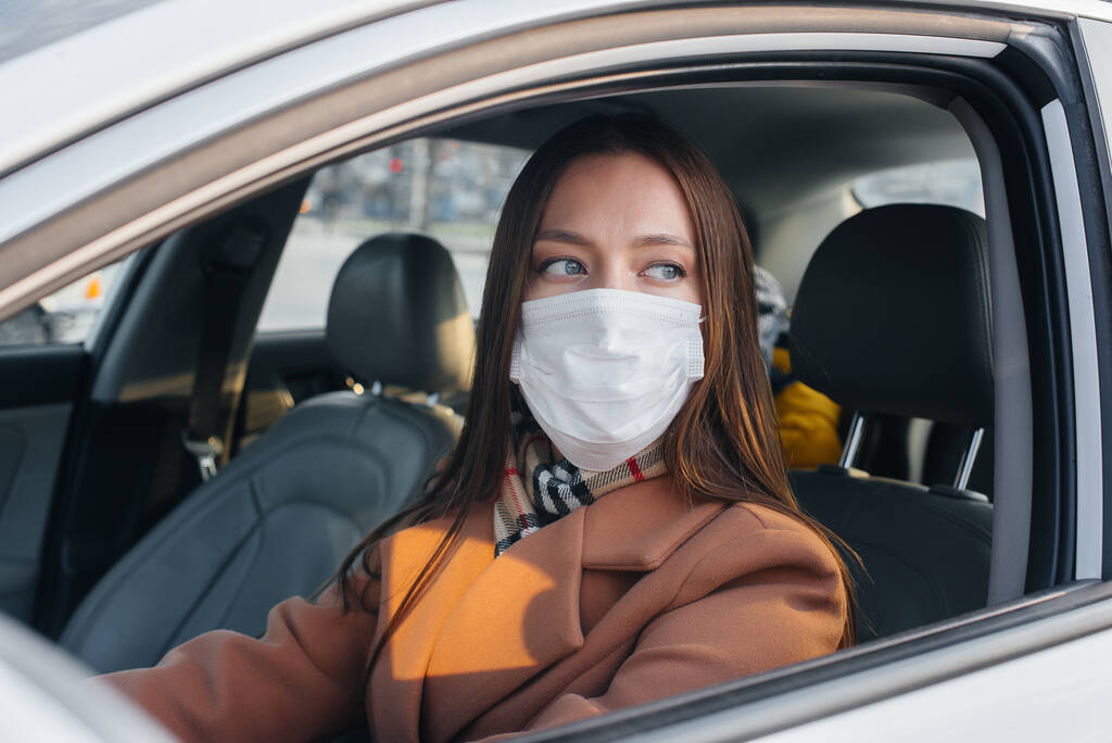 Une jeune fille est assise derrière le volant dans la voiture dans le masque pendant la pandémie mondiale et le coronavirus. Mise en quarantaine - Photo, image