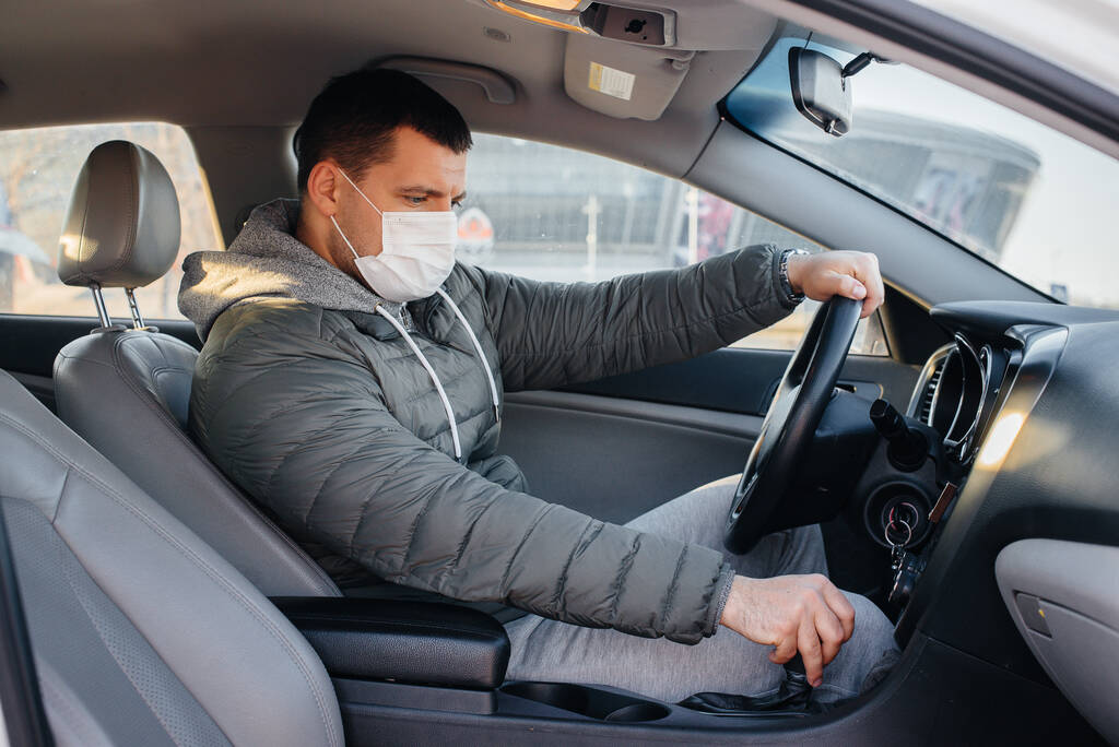 A young man sits behind the wheel wearing a mask for personal safety while driving during a pandemic and coronavirus. Epidemic - Photo, Image