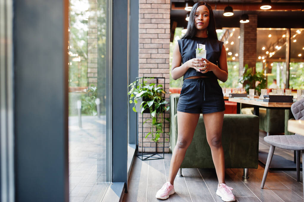 Donna afroamericana femminista alla moda vestita in t-shirt e pantaloncini neri, posata al ristorante con vetro limonata. - Foto, immagini