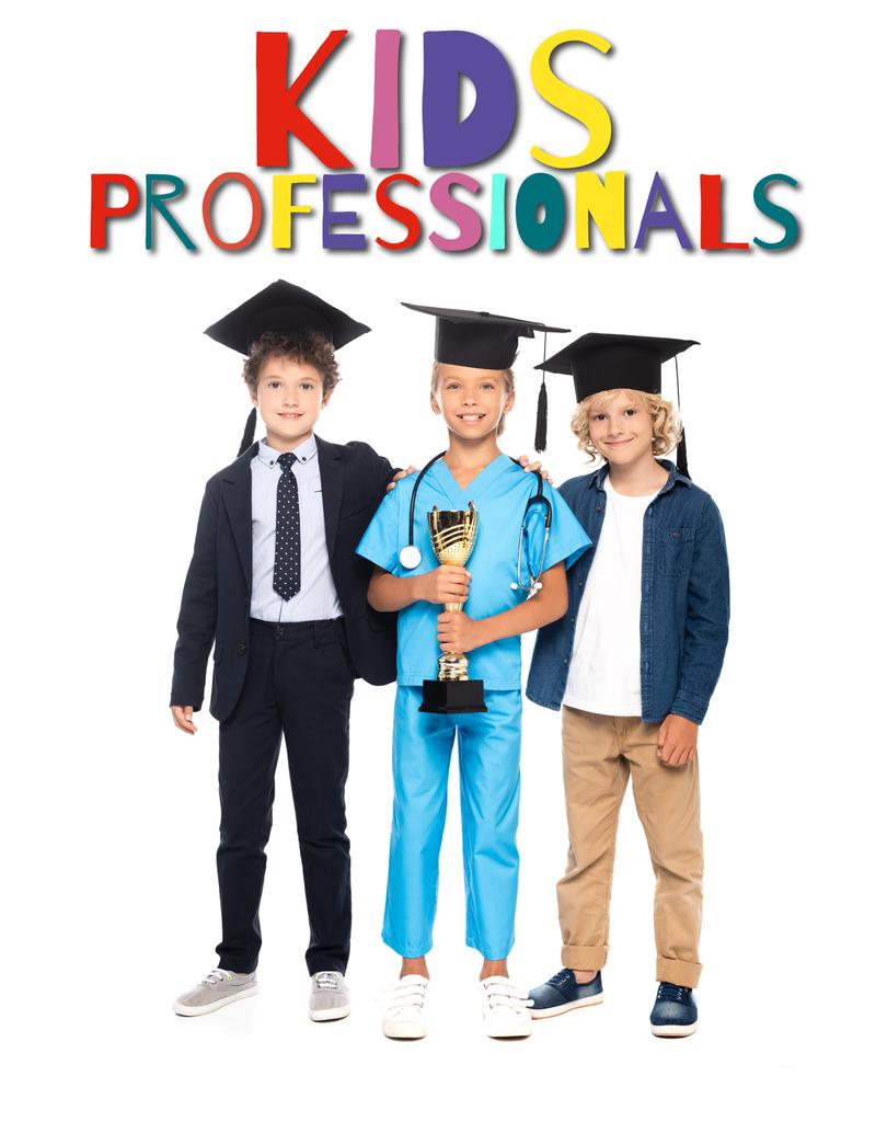 bambini in berretti di laurea vestiti in costumi di diverse professioni che detengono trofeo d'oro vicino a bambini professionisti lettering su bianco  - Foto, immagini