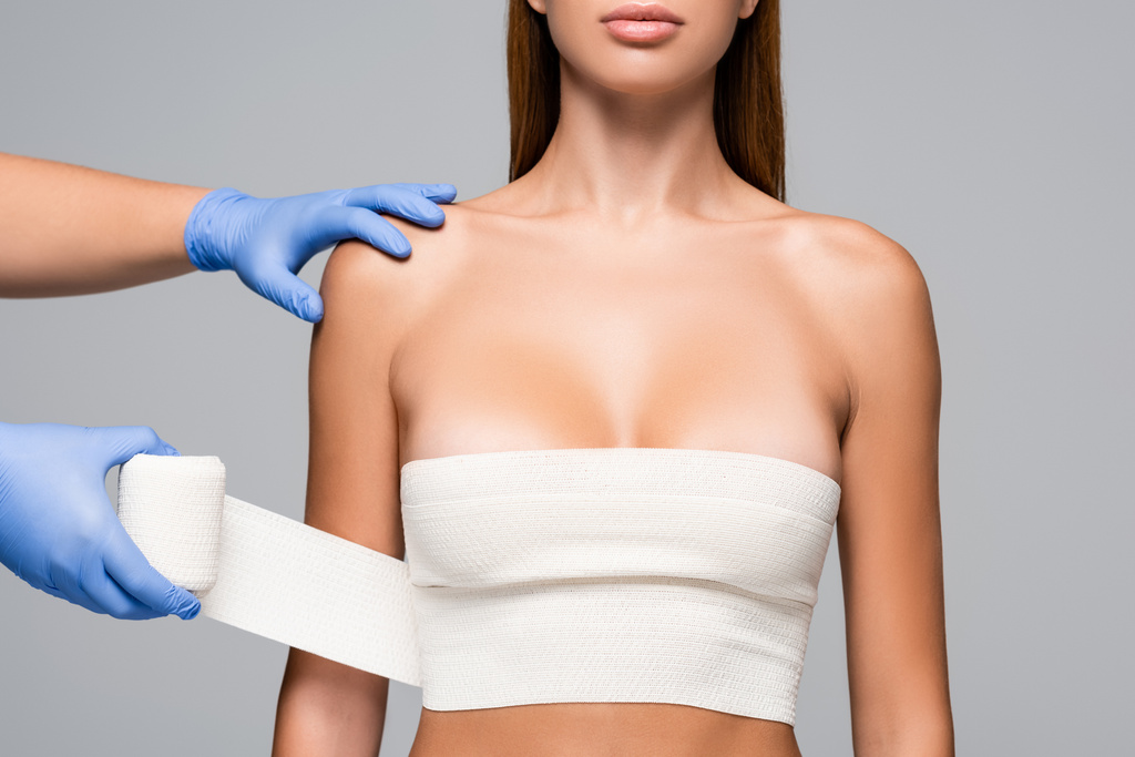Vista recortada del médico en guantes de látex envolviendo pecho de mujer desnuda con vendaje elástico aislado en gris  - Foto, Imagen