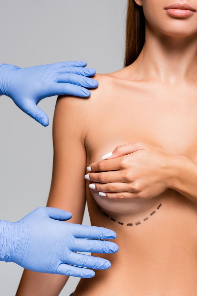Обрезанный вид пластического хирурга в латексных перчатках касающихся обнаженной женщины с отметинами на груди, изолированными на серой  - Фото, изображение