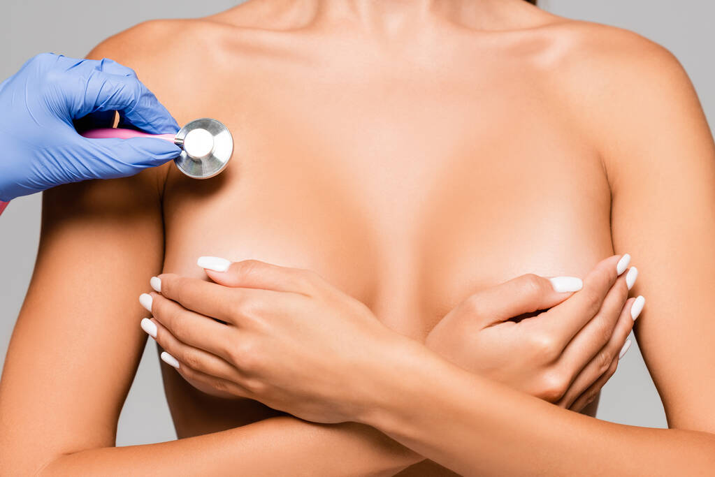 Oříznutý pohled na nahou ženu pokrývající poprsí v blízkosti lékaře se stetoskopem izolované na šedé  - Fotografie, Obrázek