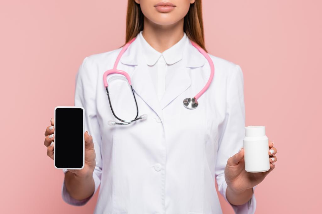 ピンクの上に隔離された錠剤と空白の画面と瓶でスマートフォンを保持医師の作物ビュー - 写真・画像