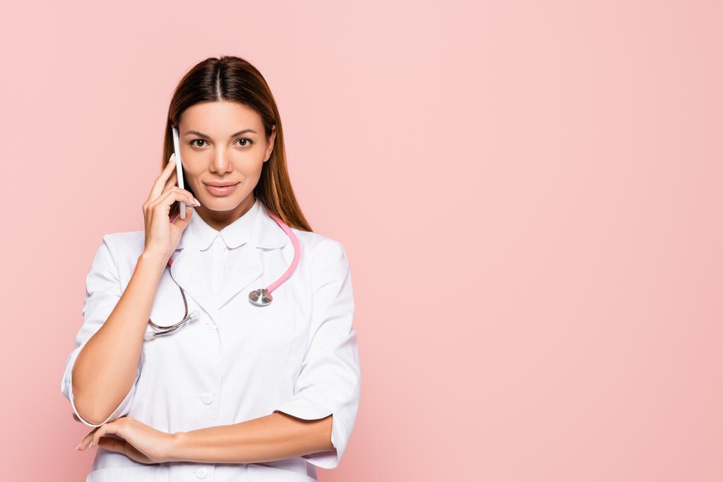 Usmívající se lékař mluví na smartphone při pohledu na kameru izolované na růžové - Fotografie, Obrázek
