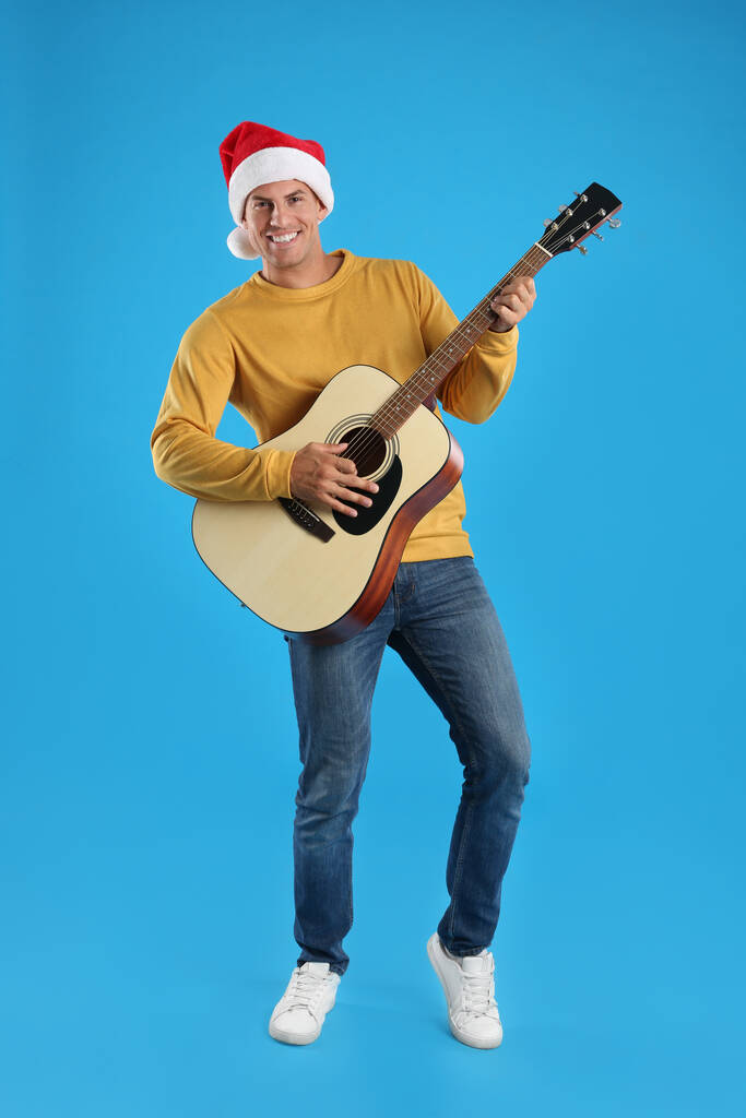 Mann mit Weihnachtsmütze spielt Akustikgitarre auf hellblauem Hintergrund. Weihnachtsmusik - Foto, Bild