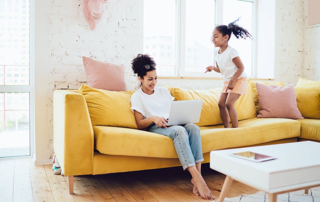 Етнічна дочка стрибає біля матері в білій сорочці і джинсах, сидячи на жовтому дивані і працюючи на ноутбуці вдома
 - Фото, зображення