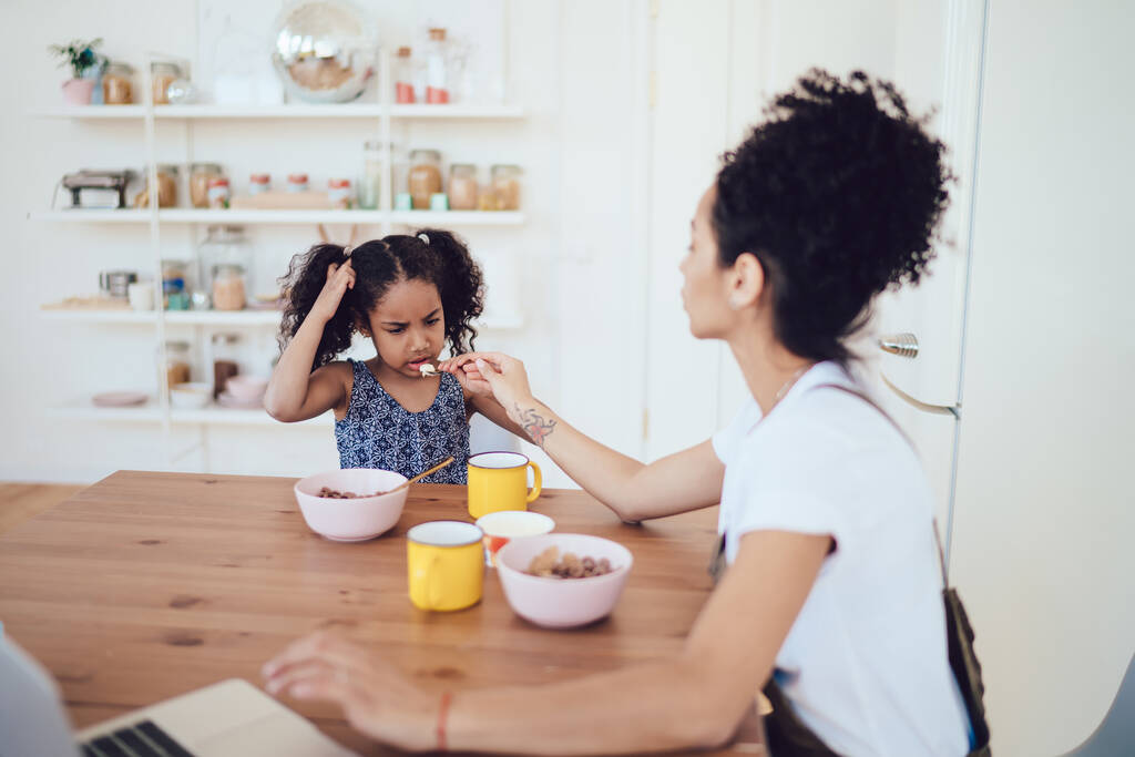 Curly African American mother in home clothes sitting at table and trying to feed capricious daughter with healthy breakfast food in morning - Photo, Image