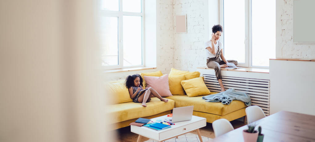 Afroamerikanisch lockiges Mädchen sitzt auf gelbem Sofa und spielt auf Tablet, während junge Mutter auf dem heimischen Fensterbrett im hellen Zimmer mit dem Handy telefoniert - Foto, Bild