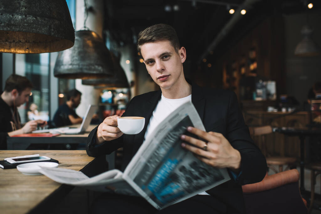 İyi giyimli öğrenci, elinde gazete ve bir fincan sıcak içecekle koyu renkli bir kafede otururken kameraya bakıyor. - Fotoğraf, Görsel