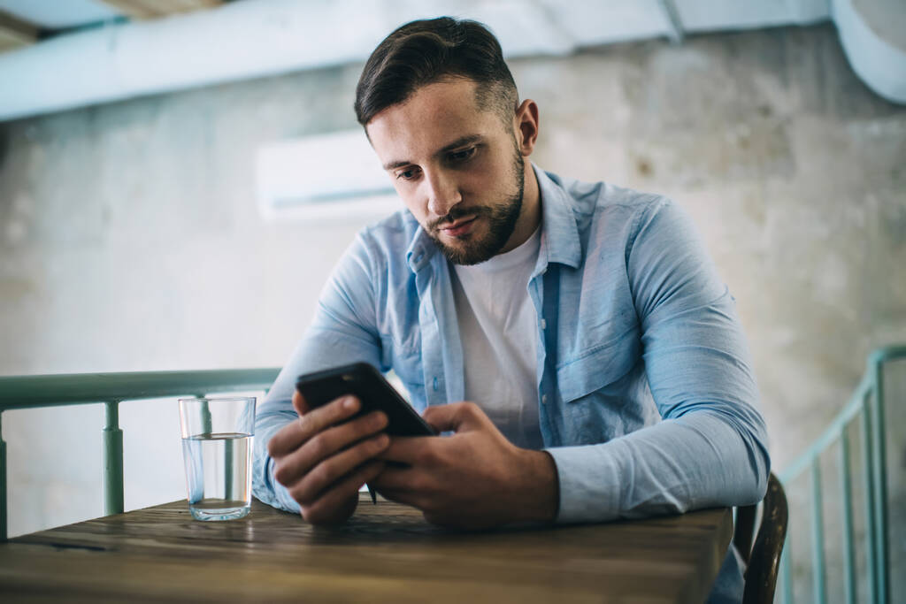 Uomo barbuto serio in abiti casual seduto sulla sedia al tavolo di legno in caffè moderno con vetro d'acqua e utilizzando smartphone - Foto, immagini