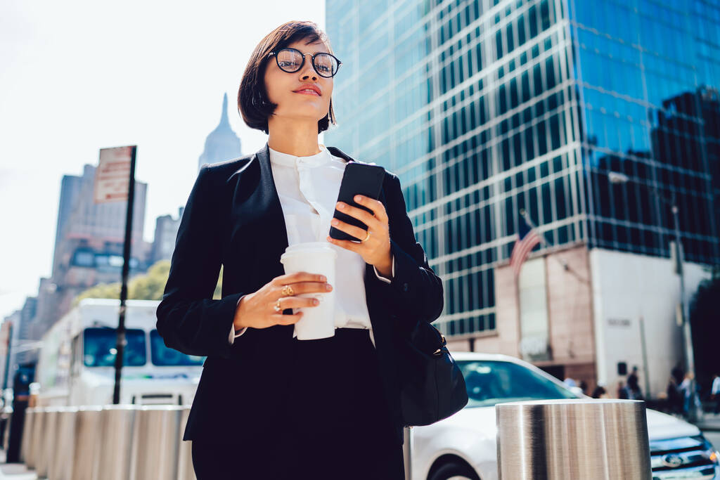 From below of content elegant businesswoman in formal clothes and eyeglasses messaging on smartphone and drinking takeaway coffee on street of New York City - Photo, Image