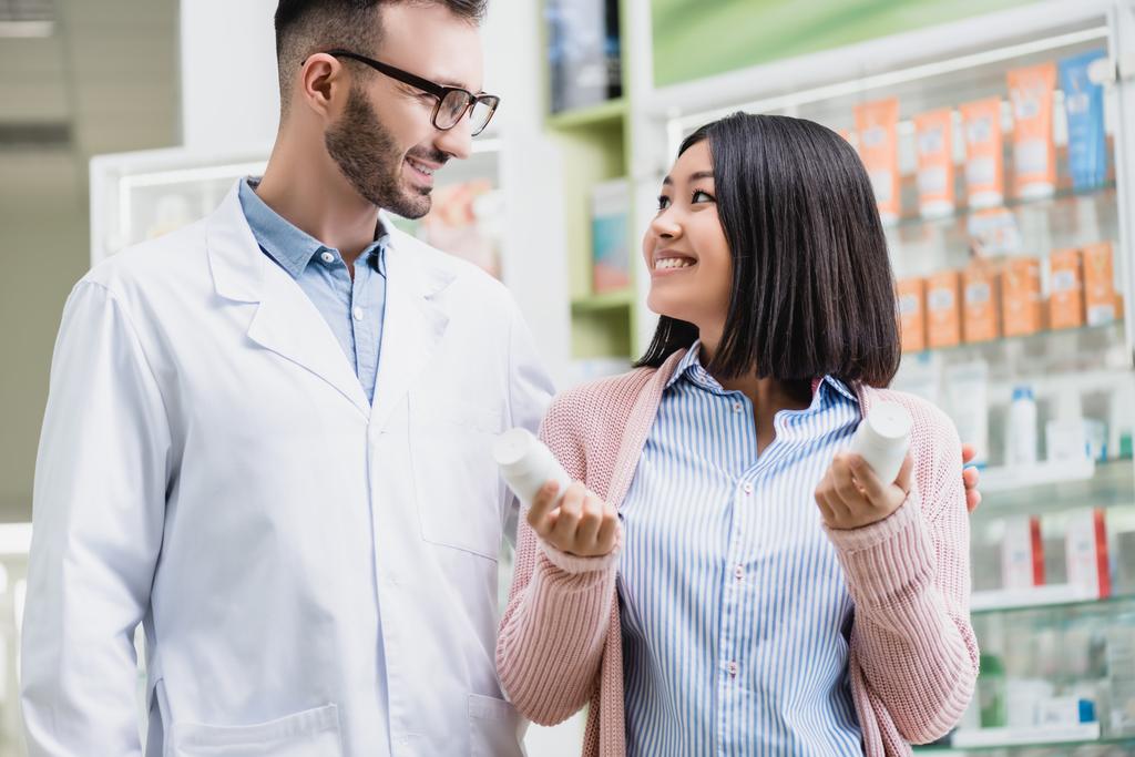 cheerful pharmacist in eyeglasses looking at asian woman holding bottles with pills in drugstore  - Photo, Image