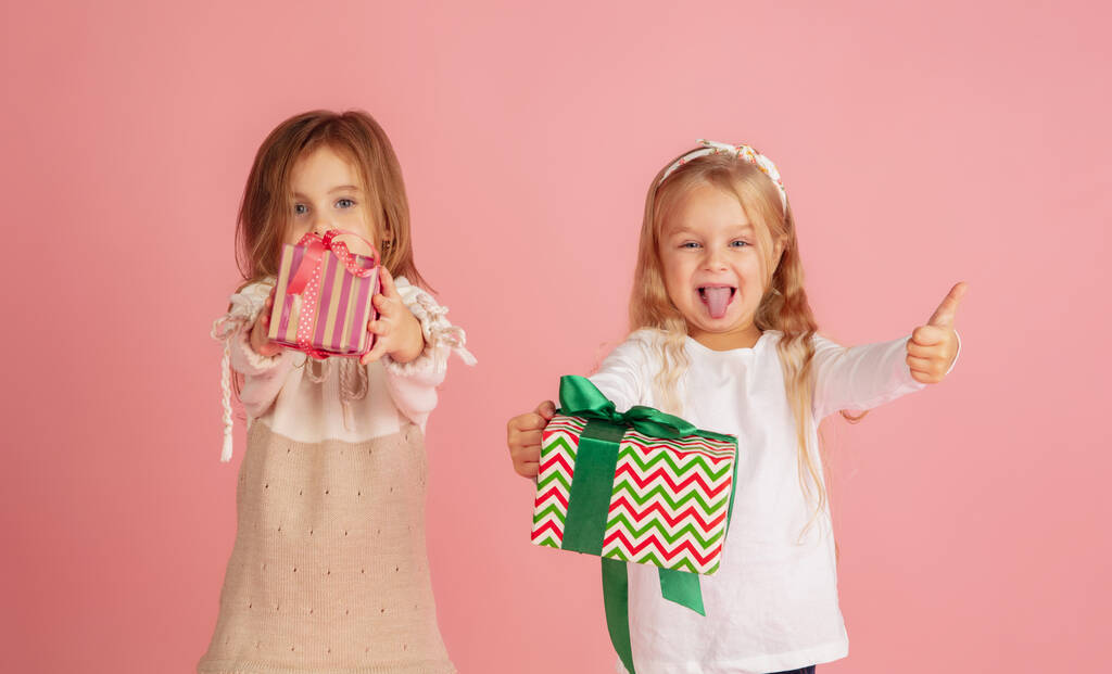 Schenken und Geschenke bekommen an Weihnachten. Zwei kleine lächelnde Kinder amüsieren sich isoliert auf rosa Studiohintergrund - Foto, Bild