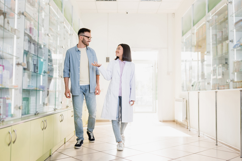 täyspitkä iloinen aasialainen farmaseutti valkoisessa takissa kävely asiakkaan kanssa apteekissa - Valokuva, kuva