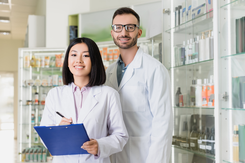 heureux pharmaciens interracial en manteaux blancs regardant caméra en pharmacie - Photo, image