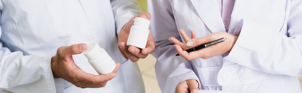 visão parcial do farmacêutico em frascos de retenção de casaco branco com medicação perto colega na farmácia, banner - Foto, Imagem