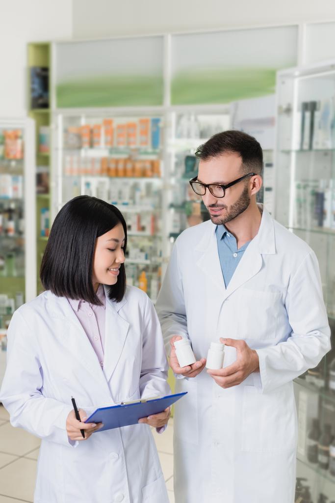 щасливий фармацевт у білому пальто тримає пляшки з ліками біля азіатського колеги в аптеці
 - Фото, зображення