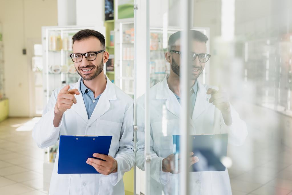 pharmacien heureux dans les lunettes tenant presse-papiers tout en pointant du doigt dans la pharmacie  - Photo, image