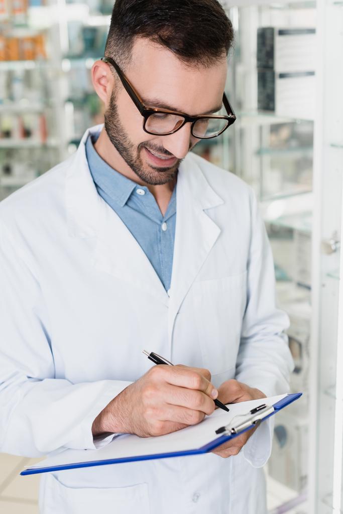 cheerful pharmacist in eyeglasses writing on clipboard in drugstore  - Photo, Image