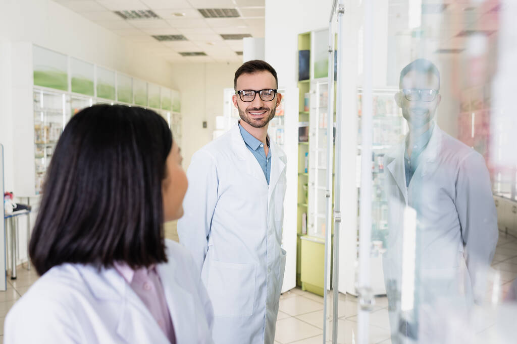 farmacêutico alegre em óculos e casaco branco olhando para morena asiático colega em primeiro plano borrado - Foto, Imagem