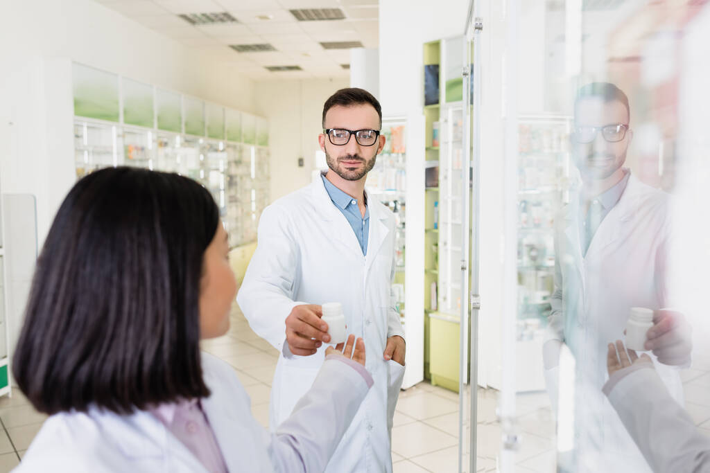 farmacêutico barbudo em óculos e casaco branco dando garrafa com pílulas para morena asiático colega em primeiro plano borrado - Foto, Imagem