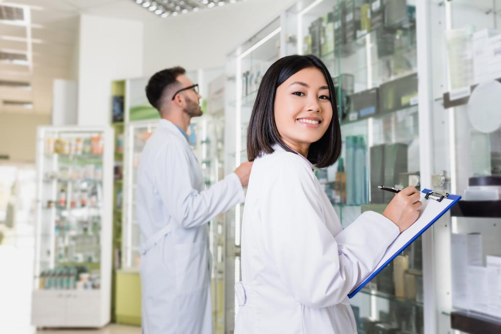 feliz asiático farmacêutico no branco casaco escrevendo na área de transferência perto colega no fundo borrado - Foto, Imagem