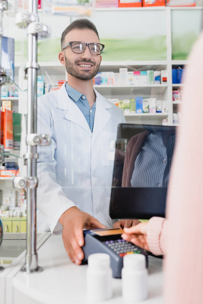 πελάτης που πληρώνει με πιστωτική κάρτα στον τερματικό σταθμό πληρωμών κοντά χαρούμενος πωλητής στο φαρμακείο  - Φωτογραφία, εικόνα