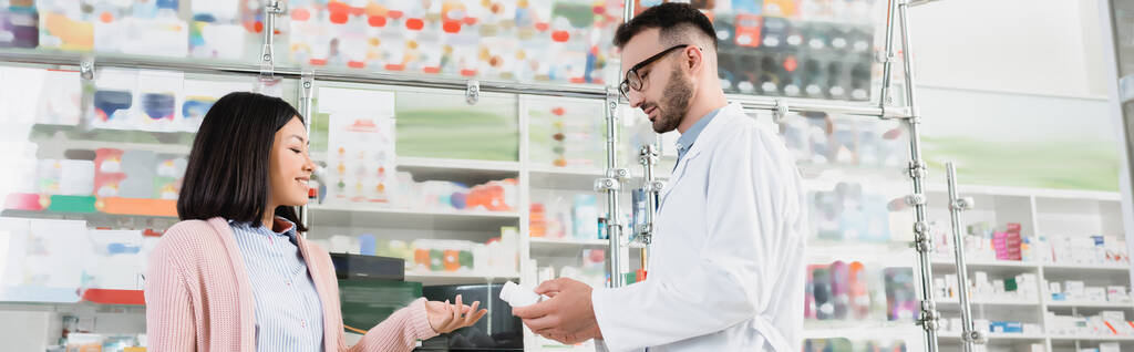 brodaty aptekarz w okularach i białym płaszczu dający butelkę z tabletkami azjatyckiemu klientowi, baner - Zdjęcie, obraz