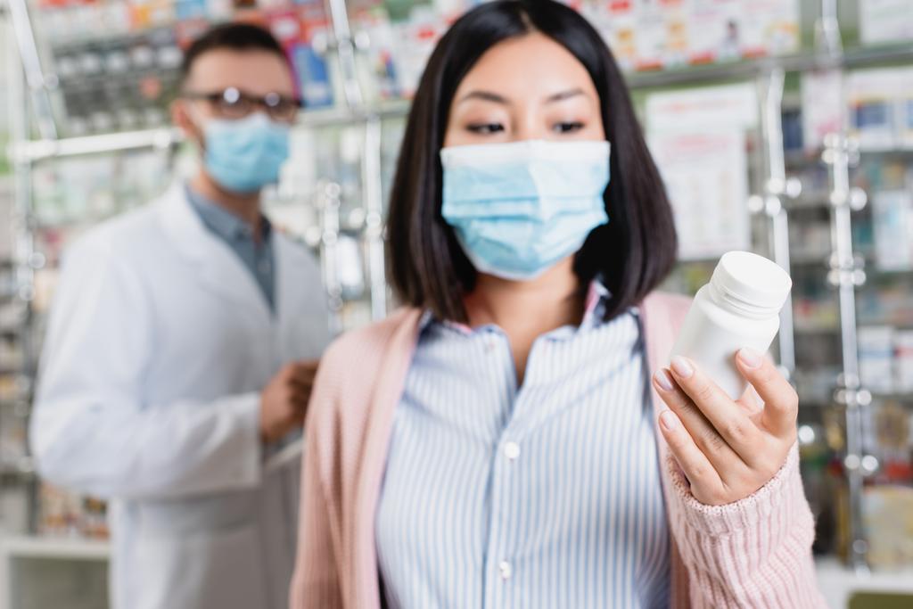 bottle with pills in hand of asian woman in medical mask near pharmacist on blurred background - Photo, Image