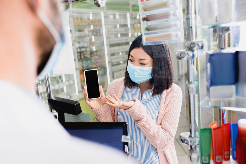 mulher asiática em máscara médica apontando com a mão enquanto mostra smartphone com tela em branco para farmacêutico em primeiro plano borrado - Foto, Imagem