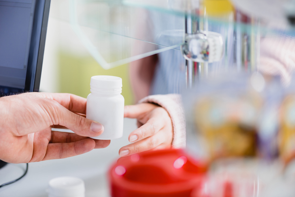 oříznutý pohled lékárníka, jak dává lahev s léky zákazníkovi poblíž monitoru počítače - Fotografie, Obrázek