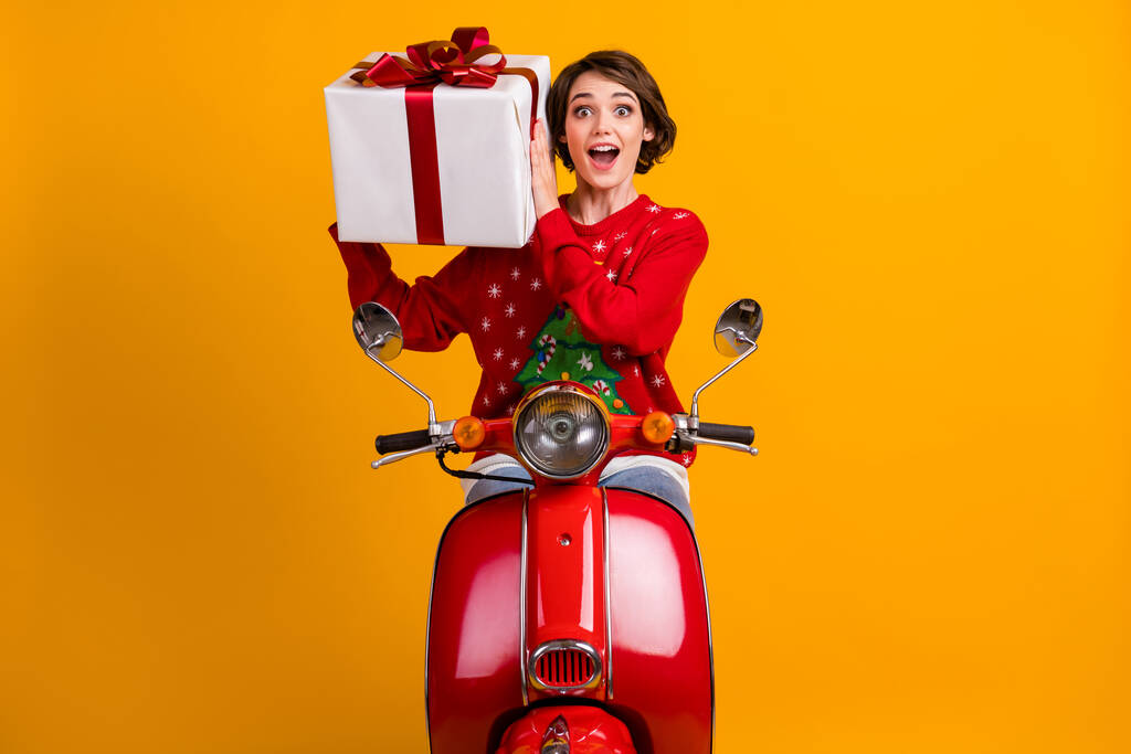 Photo de fille excitée dans l'ornement de Noël pull pull tour scooter rouge x-mas fête obtenir grande boîte cadeau curieux isolé sur fond de couleur brillante - Photo, image