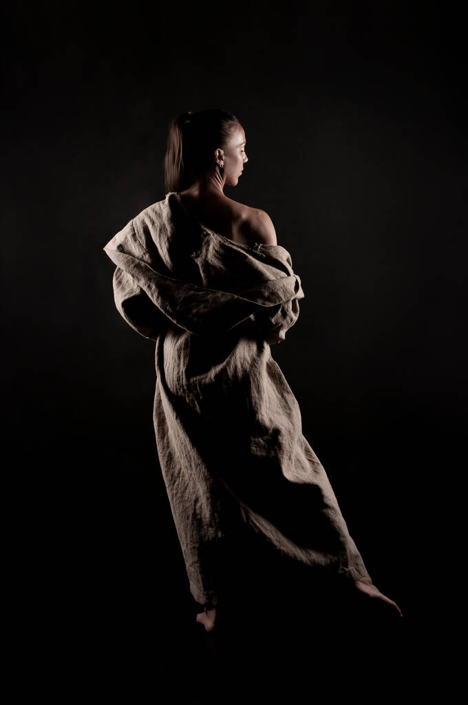 Nuori nainen karkeasta kankaasta tehdyssä sadetakissa alastomassa ruumiissaan on eristetty mustalla pohjalla. Studiokuvaus. - Valokuva, kuva