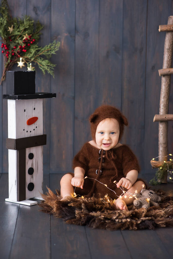 Šťastný chlapeček v hnědém, načechraném, pleteném medvědím kostýmu, sedí na furonu na dřevěné podlaze a hraje si s jasným vánočním věncem světel. Blízko je sněhulák. Novoroční přání. Kopírovat prostor  - Fotografie, Obrázek