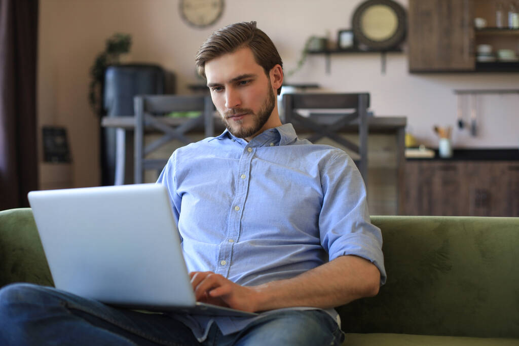 Konsantre olmuş genç serbest çalışan işadamı dizüstü bilgisayarla kanepede oturuyor, uzaktan evde çalışıyor. - Fotoğraf, Görsel